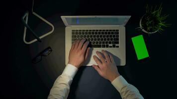 Hands of a man in a white shirt are typing on a laptop keyboard while sitting at a table at night. Overhead shot. video