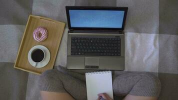 Overhead shot, creative girl draws a graph in a notebook sitting on the bed next to a laptop and a cup of coffee with a donut video