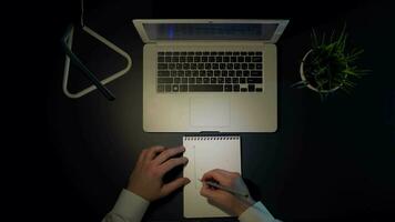 uomo nel bianca camicia si siede a il tavolo e disegna un' grafico di redditi nel un' taccuino mentre seduta a un' il computer portatile nel il sera alto tiro video