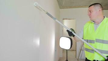en man målarfärger en vägg i en rum vit. reparera arbete i de rum av de hus. de målare rullar vit måla på de vägg med en vält. video