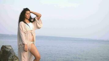 beautiful girl model with long hair and in a beach tunic is standing on stones against the background of the sea and posing. slow motion video