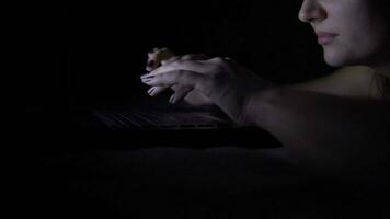 Attractive young girl typing on a laptop keyboard while lying on a bed at night. video