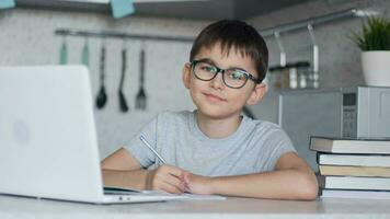 Attractive child in glasses teaches lessons while at home sitting in the kitchen at the table using a laptop, books, notebook and looking at the camera with a smile. Left camera movement video