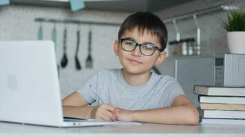 Attractive child in glasses teaches lessons while at home sitting in the kitchen at the table using a laptop, books, notebook and looking at the camera with a smile. Close-up video