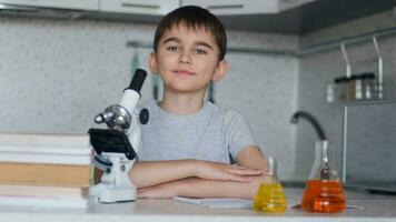 Portrait of a smart attractive schoolboy who sits at a desk at home next to a microscope and flasks with liquid video