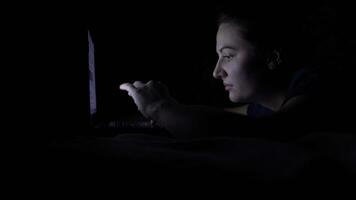 An attractive young happy woman uses a mobile computer while lying on a bed at night. Isolated light 4K video