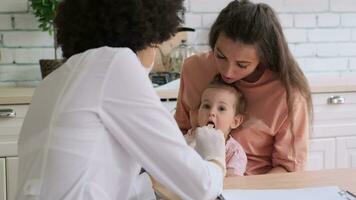 Afro american woman doctor in mask looking at baby's throat at home while sitting at the table at home next to mom. Family Doctor, Patient Support, Help at Home, Caring for the Sick. video