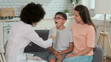 Afro american woman doctor in mask looks at sick child's throat while examining at home next to mom. Family Doctor, Patient Support, Help at Home, Caring for the Sick. video