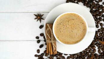 Cup of fresh coffee, cinnamon sticks and scattered coffee beans on a white wooden background. Coffee day. Close-up. Top view. Place for text. Selective focus. photo