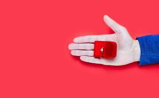 Male hand holding a gift box with engagement ring on red background. Close-up. Top view. Place for text. photo