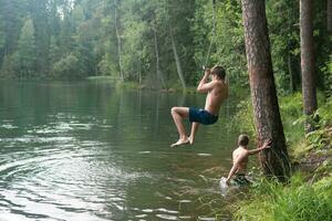 Niños saltos dentro el agua utilizando un tarzán columpio mientras nadando en un bosque lago foto