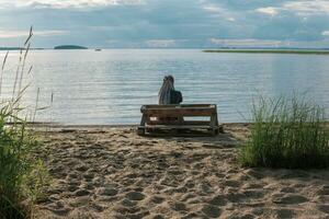 woman traveler with de braids hairstyle sitting on a bench made of old pallets on a sandy shore of a vast lake photo