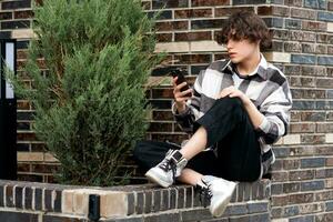 young man reads something on the phone while sitting on the brick curb photo