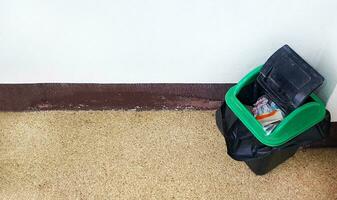 Top view of black and green bin putting on corner on floor with white wall background and copy space. Flat lay of plastic garbage on ground. photo