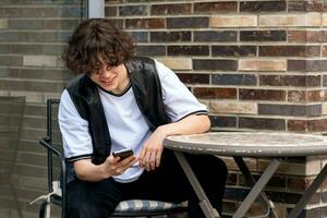 joven hombre leyendo gracioso mensaje en teléfono mientras sentado a un al aire libre cafetería mesa foto