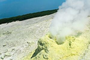 cone of sulfur deposits around a fumarole in a solfataric field illuminated by the sun against a stormy landscape in the distance photo