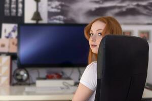 young woman sitting at a table in front of a computer monitor turning around photo