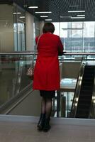 joven mujer en un rojo Saco soportes con su espalda a el espectador en el vestíbulo de un público edificio foto