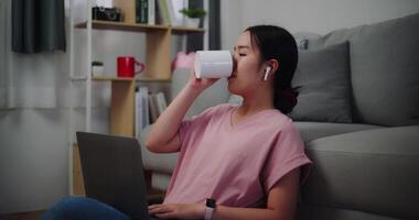 Footage of Young woman sitting on the floor leaning against a sofa holding coffee to drinking while working on laptop at home office. video