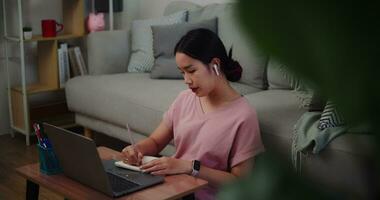 metraggio di giovane donna seduta su il pavimento pendente contro un' divano Lavorando con un' il computer portatile e prendere Appunti nel un' taccuino a casa ufficio. video