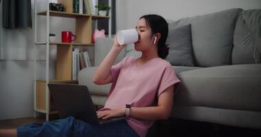 metraggio di giovane donna seduta su il pavimento pendente contro un' divano Tenere caffè per potabile mentre Lavorando su il computer portatile a casa ufficio. video