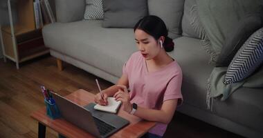 Footage of Young woman sitting on the floor leaning against a sofa working with a laptop and take notes in a notebook at home office. video