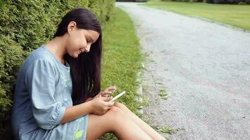 een meisje met lang zwart haar- zit Aan de gras in de park en toepassingen een tablet en glimlacht Aan een zonnig dag video
