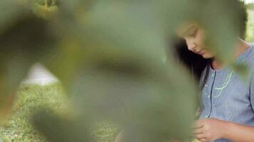 retrato. un hermosa pequeño niña con largo negro pelo y en un azul vestir leyendo un libro sentado debajo un árbol en el césped trae el libro a su cofre y Sueños de sonriente en un soleado verano día video