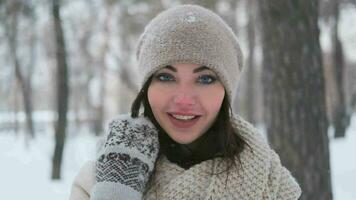 retrato de un hermosa niña con azul ojos y oscuro pelo en mitones posando en un invierno parque sonriente y corriendo un mano mediante su cabello.lento movimiento video