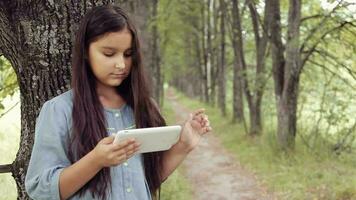 retrato. un hermosa niña con largo negro pelo en un vestir es en pie por un árbol en el naturaleza utilizando un tableta y mirando a alguna cosa interesante en eso y riendo video