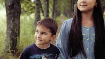 Porträt. glücklich schön Mutter mit Tochter und Sohn spazieren gehen durch das Park halten Hände lächelnd und genießen Natur auf ein Sommer- sonnig Tag während das Tochter zeigt an etwas interessant video