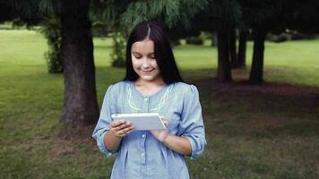 Portrait. A beautiful girl with long black hair in a dress is walking in nature using a tablet and looking at something interesting in it and smiling video