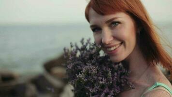 retrato. de cerca. lento movimiento. hermosa joven sexy niña con largo rojo pelo en un turquesa vestir soportes con un hermosa sonrisa con flores en el antecedentes de el mar video