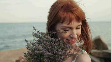 retrato. fechar-se. lento movimento. lindo jovem sexy menina com grandes vermelho cabelo dentro uma turquesa vestir positivamente posando em a mar com flores dentro mãos video