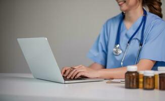 Young Asian doctor in a white medical uniform with a stethoscope using a lab computer and tablet to organize and classify medicines in preparation for administration to patients. photo