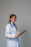 retrato de un sonriente hembra médico participación un portapapeles, vistiendo un médico Saco y estetoscopio, gris antecedentes. foto