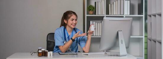 Young beautiful doctor holding pill bottle explaining details about medicine via laptop online at clinic photo