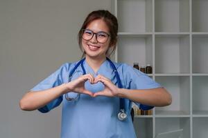 Beautiful asian woman wearing doctor uniform and stethoscope smiling in love doing heart symbol shape with hands. romantic concept. photo