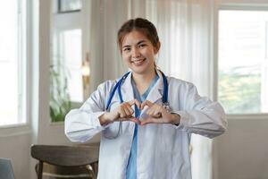 hermosa asiático mujer vistiendo médico uniforme y estetoscopio sonriente en amor haciendo corazón símbolo forma con manos. romántico concepto. foto