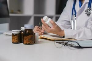 Asian female medic doctor gp therapist holding pills bottle in hand writing medical prescription sitting at work desk prescribing pharmacy medicine concept. Closeup view photo