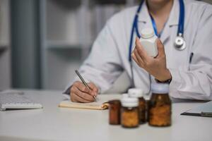 Asian female medic doctor gp therapist holding pills bottle in hand writing medical prescription sitting at work desk prescribing pharmacy medicine concept. Closeup view photo