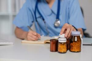 Asian female doctor, GP therapist holding pill bottle in hand, writing a prescription, sitting at prescription table, pharmaceutical concept. Closeup view. photo