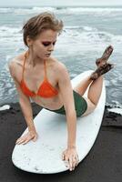Female surfer lies on front of surfboard, arching her back and crossing legs while leaning on hands photo