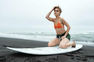 Woman surfer posing on knees on surfboard, looking at camera on beach on background of ocean waves photo