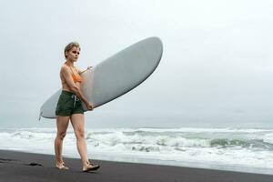 Woman surfer walks on black sand beach carrying white surfboard against backdrop of sea waves photo