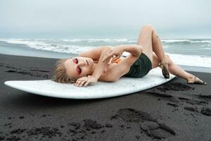 hembra tablista acostado en blanco tabla de surf en negro arenoso playa. Deportes Moda modelo con cerrado ojos foto