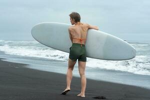 Rear view of female surfer walking on sandy beach carrying surfboard on background of ocean waves photo