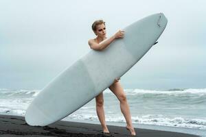 Female surfer on sandy beach holding white surfboard, standing behind it. Woman sports fashion model photo