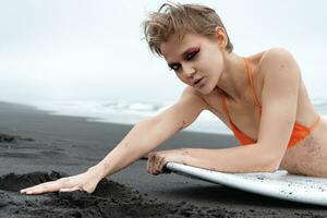 Portrait of blonde surfer woman with bright makeup lying in front of surfboard on sandy beach photo