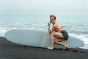 Female sports fashion model holding surfboard and sits in front of it on background of ocean waves photo